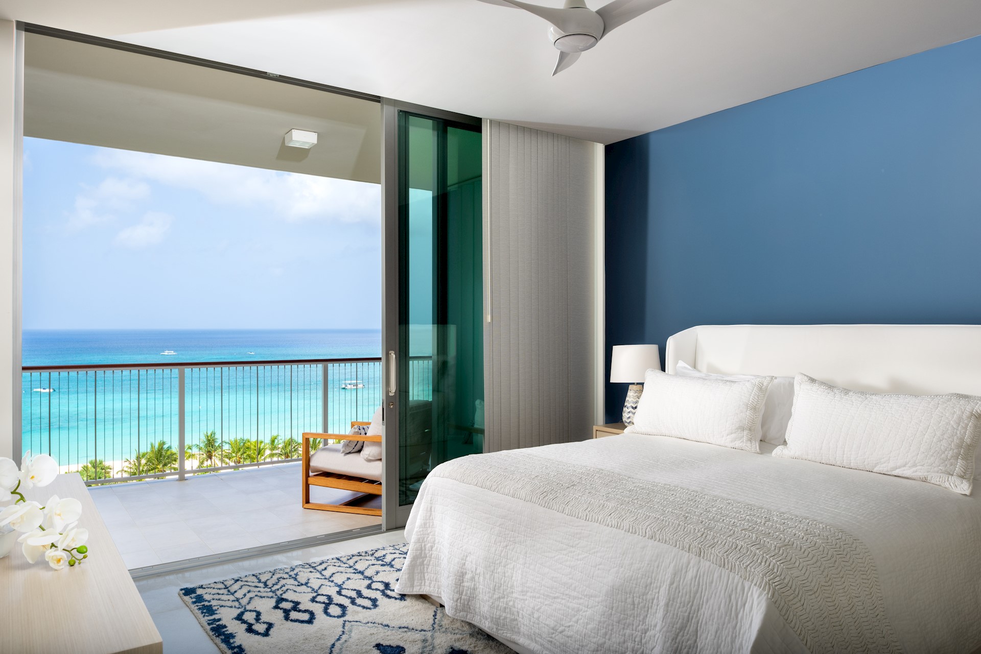 Bedroom with patio doors open and overlooking the caribbean sea. The bed sits in front of a dark blue wall and has white pillows and a white duvet.