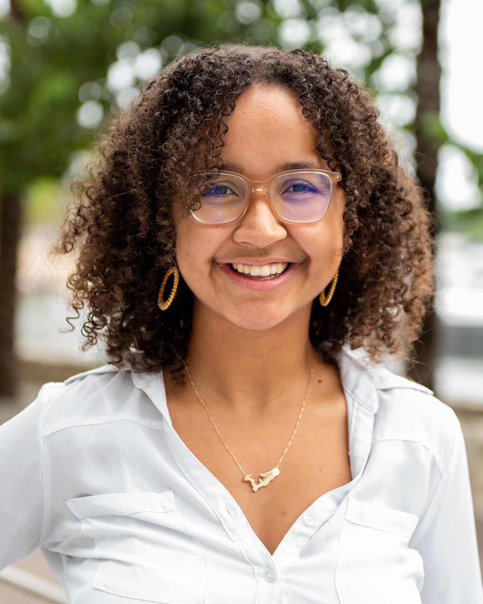 Brown skinned biracial woman with curly brown hair wearing glasses and a white shirt smiles at the camera