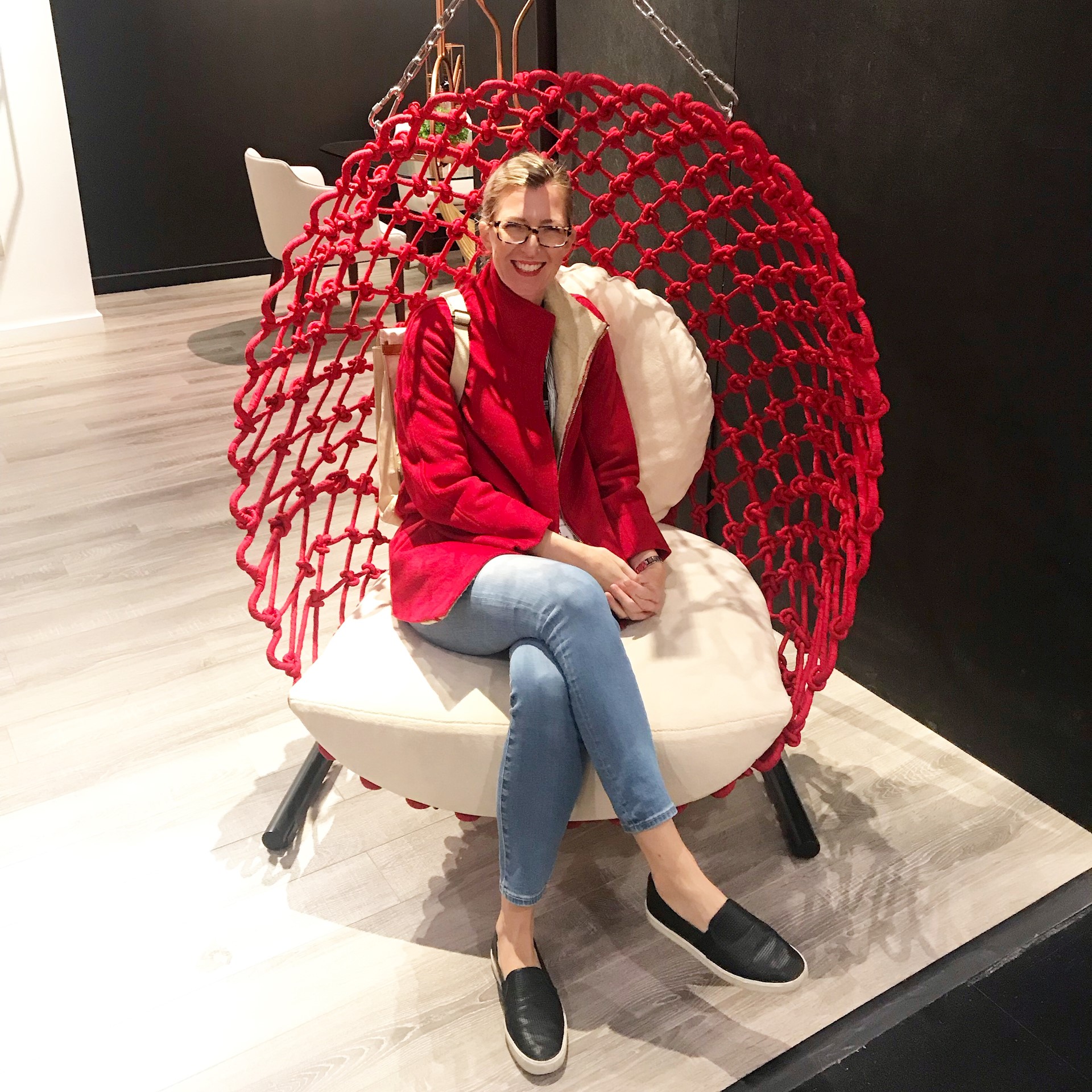 Blonde Caucasian women sitting in a red chair wearing a red jacket and blue jeans smiles at the camera