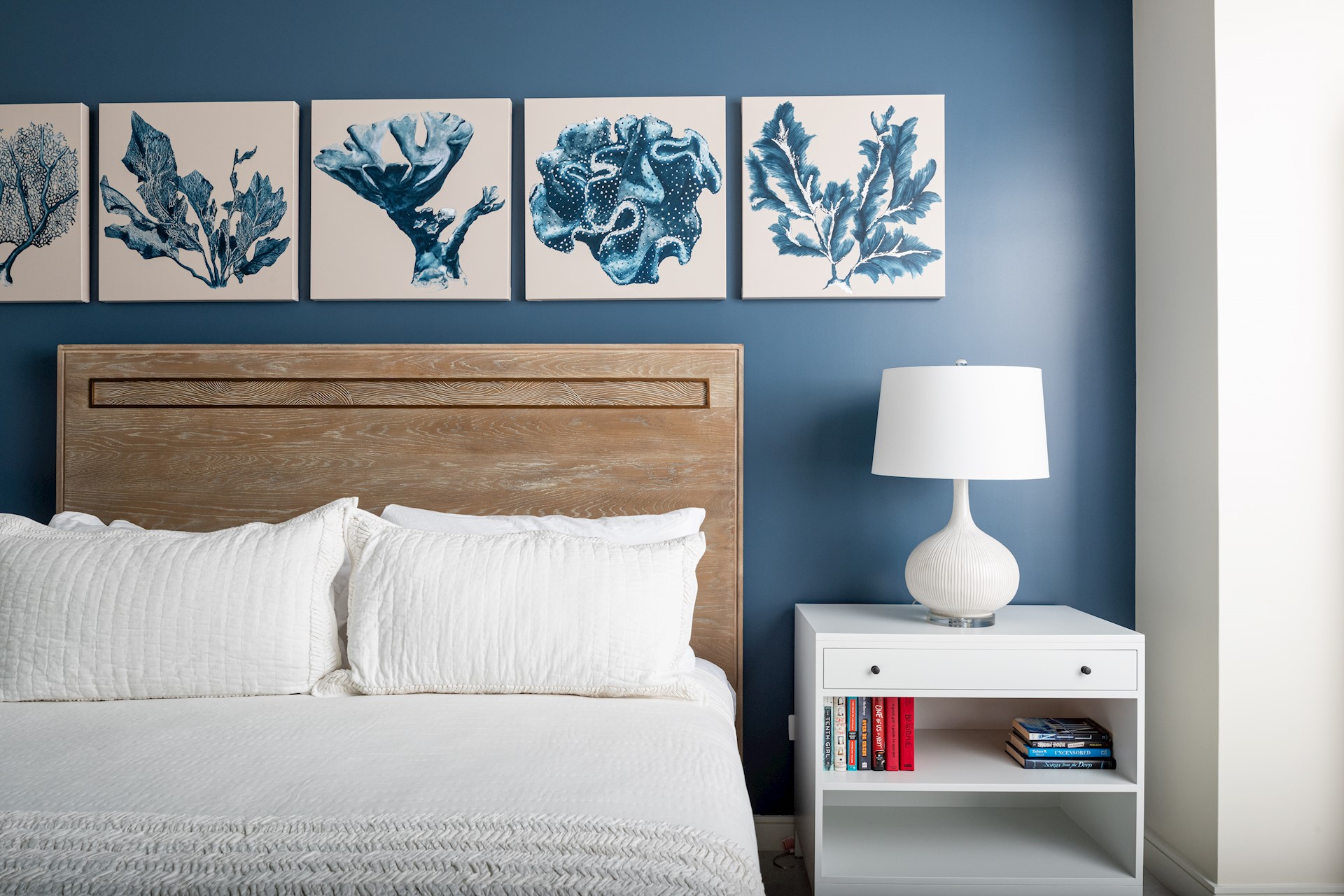 A bed with a white duvet and white pillows and a wooden headboard next to a white sidetable and white lap. The furniture is in front of a dark blue wall with five paintings on it,